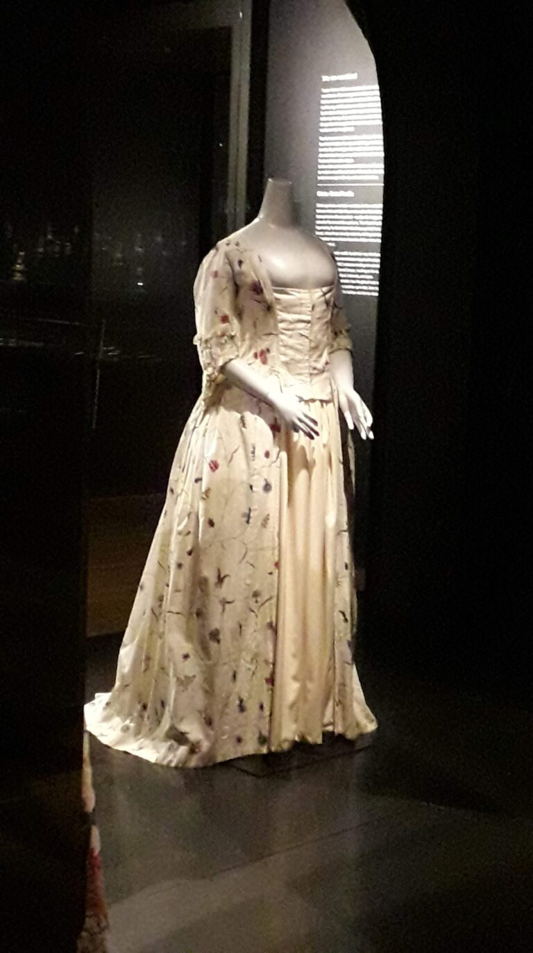 1700s cream and embroidered dress in a museum setting with dark/ black background