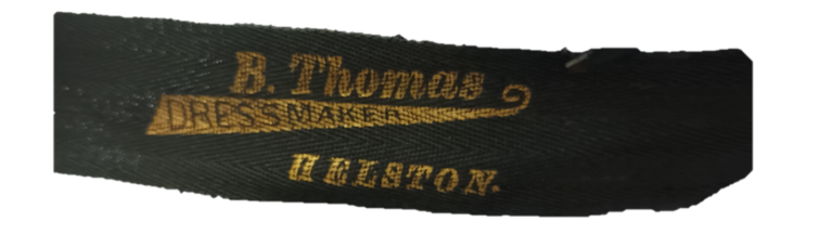 Black ribbon with gold text reading B. Thomas, Dress Maker, Helston