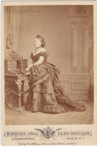 A sepia photograph of a victorian woman wearing an off the shoulder dress where the top layer of the skirt sweeps back to add volume behind and train - this is called a Bustle