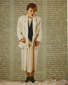 Newspaper pattern background with a woman wearing a baggy white suit jacket and matching 3/4 length skirt with padded shoulder pads.