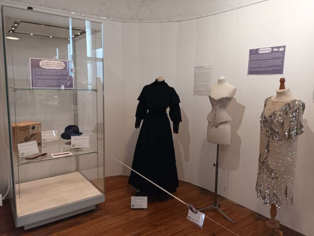 Ideal Body Shape section of Stories Told in Stitches Exhibition. A glass case on the left shows a hat box, bowler hat and brush with interpretation notes. The middle and right feature manequins wearing a 1840s mouring dress, a 1900s s-bend corset, and a 1920s flapper dress.