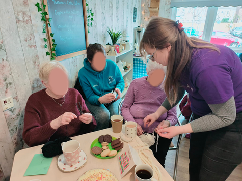 Molly is seen working with 3 seated older ladies attending spinning. The table features hot drinks and biscuits.