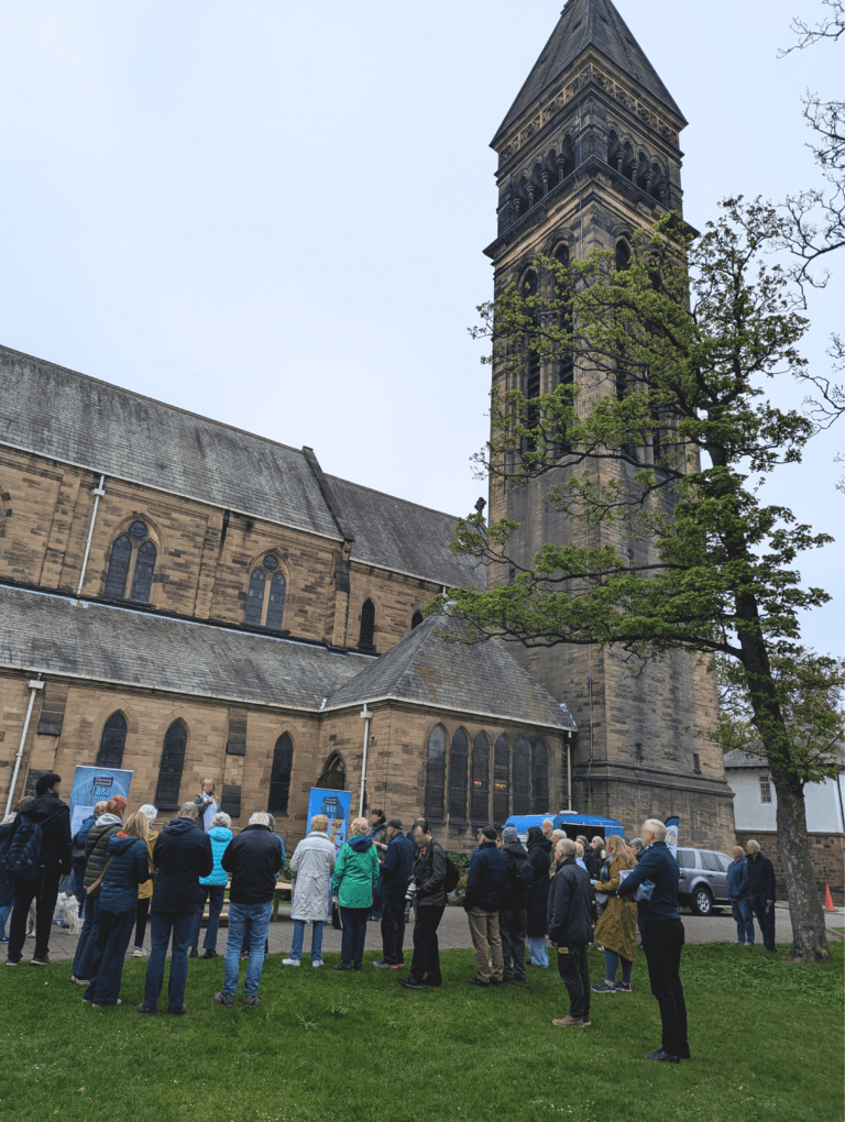 Group of about 40 people seen gathered in front of St George's, Jesmond listerning to a woman speaking on stage. Coffee van can be seen.