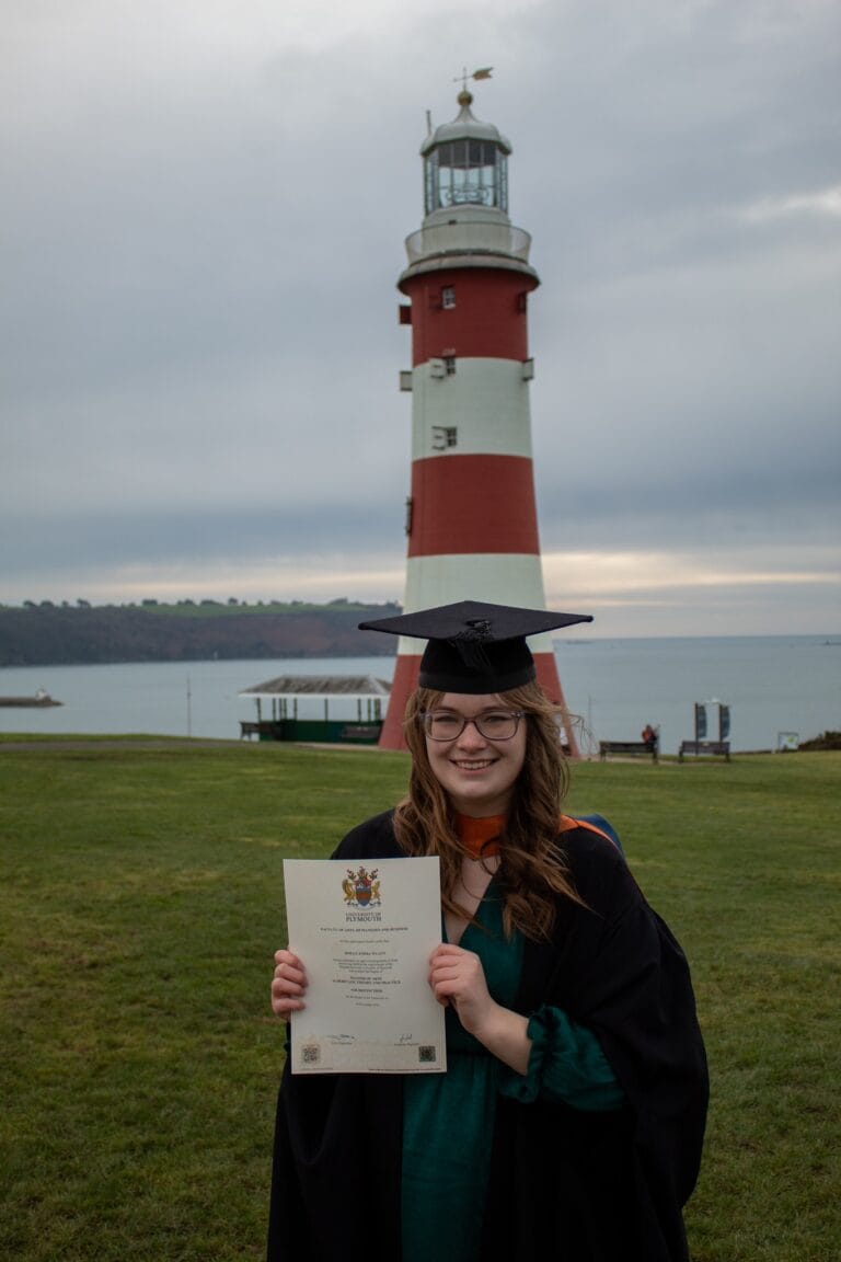 Molly posed with MA Heritage Theory & Practice certificate in graduation robes in front of Plymouth Sound lighthouse.