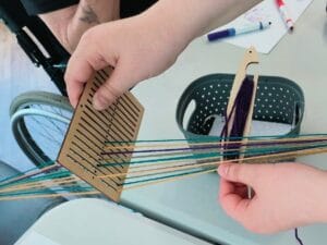 A heddleboard (a wooden board with small slots and holes) with thread in multicolours.