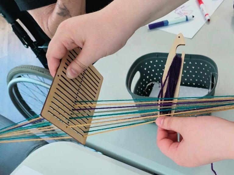A heddleboard (a wooden board with small slots and holes) with thread in multicolours.