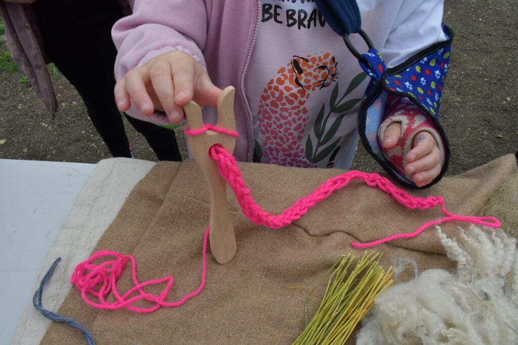A young girl with an arm in a sling holds a wooden lucet fork with neon pink wool and a partly made bracelet