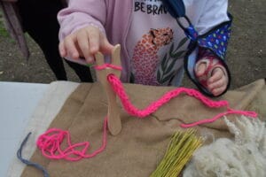 A young girl with an arm in a sling holds a wooden lucet fork with neon pink wool and a partly made bracelet