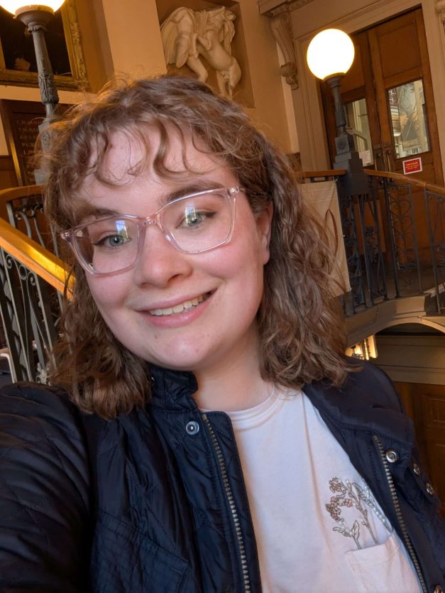 Molly is a white girl in her mid 20s with curly hair and fringe. She's pictured in front of a stately staircase at the Lit & Phil library.